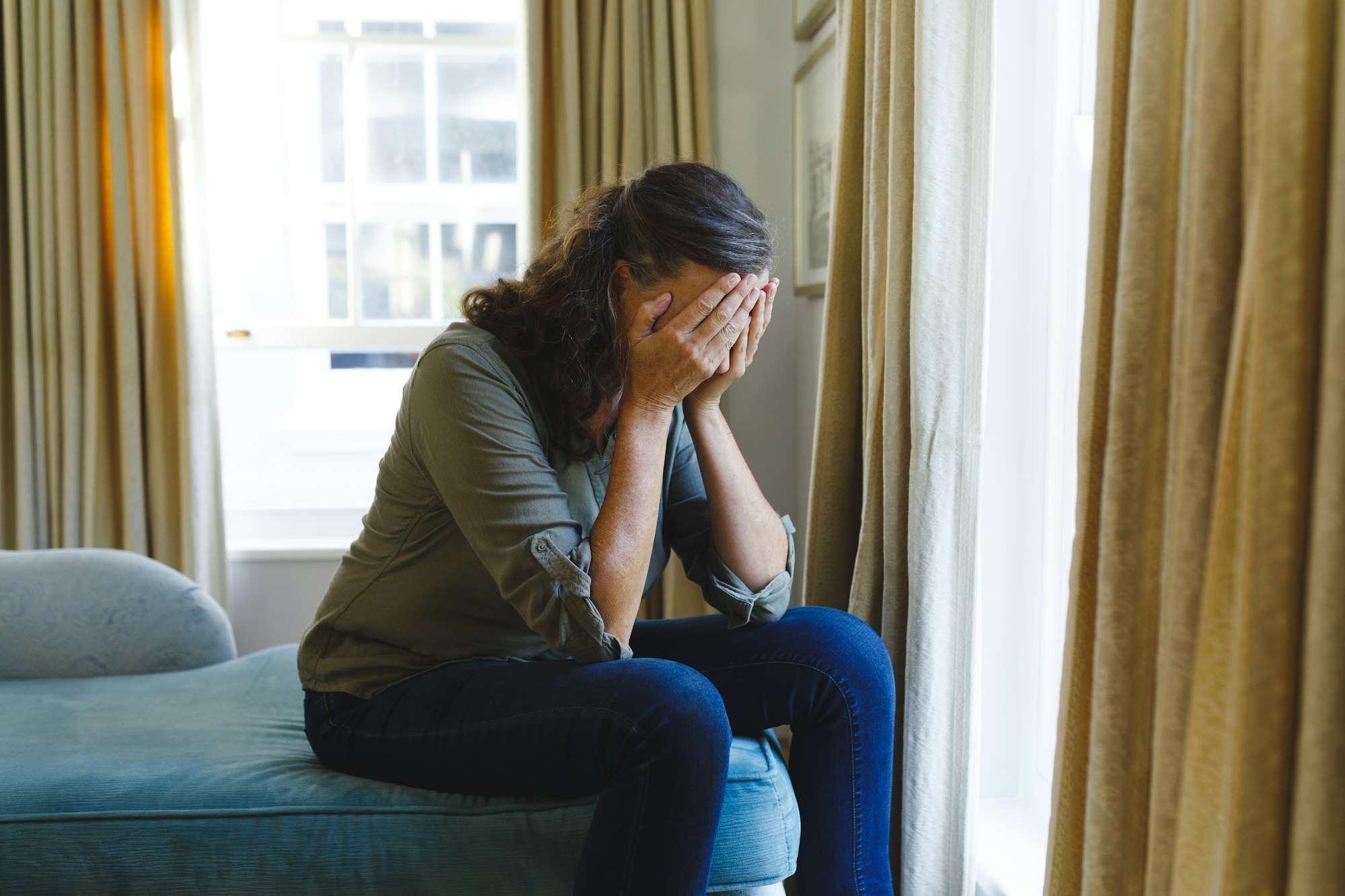 Stressed senior caucasian woman sitting on sofa by window covering face